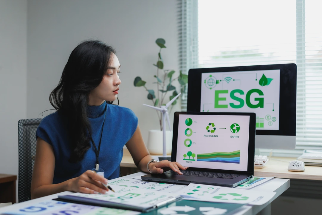 A young adult woman with dark, wavy hair, wearing a blue top, sits at a desk and studies environmental, social, and governance (ESG) data.